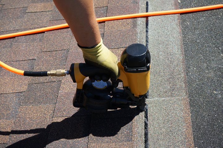 Construction worker installing asphalt roof shingles with a yellow pneumatic nail gun, wearing protective gloves, connected to an orange air hose on a residential rooftop.
