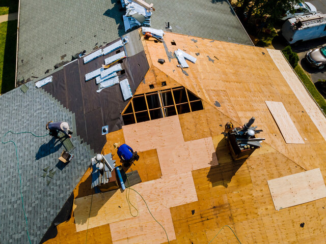 Aerial view of roof replacement in progress with workers installing shingles and underlayment over new plywood decking on a residential house. Roofing tools, materials, and safety equipment visible during home roof construction.